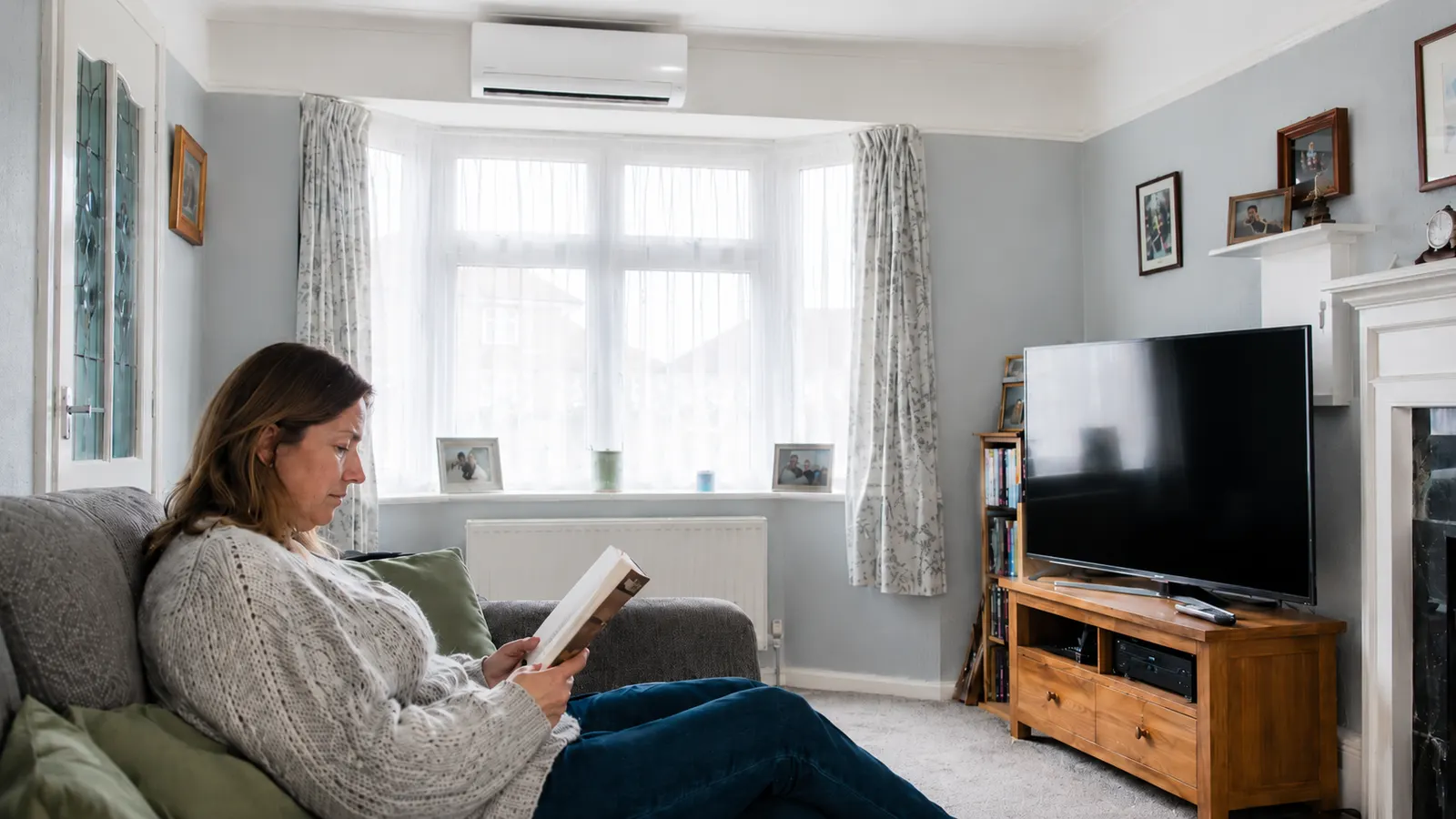 Woman reading on a sofa in a UK living room with a wall-mounted air conditioning unit above the bay window
